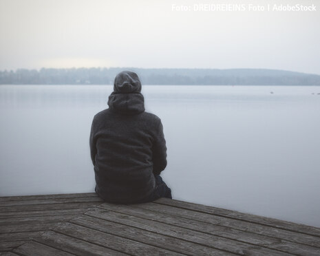 Eine Person sitzt mit dem Rücken zum Betrachter auf einem Steg und blickt auf einen See. Die Person trägt dunkle Kleidung und eine dunkle Mütze.